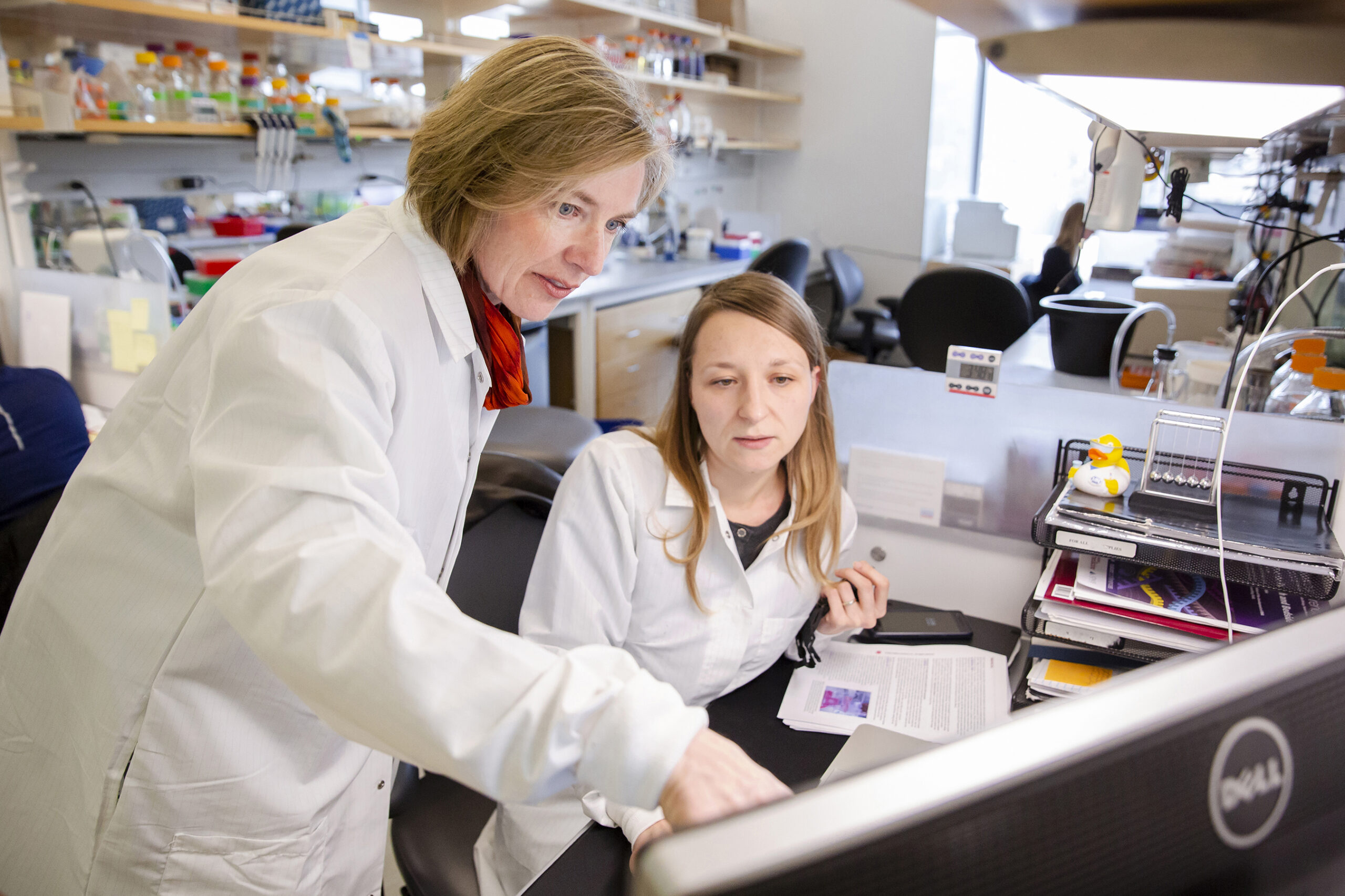 Jennifer Doudna, in a white lab coat, points at computer screen while speaking to a student in her biochemistry lab at the Innovative Genomics Institute